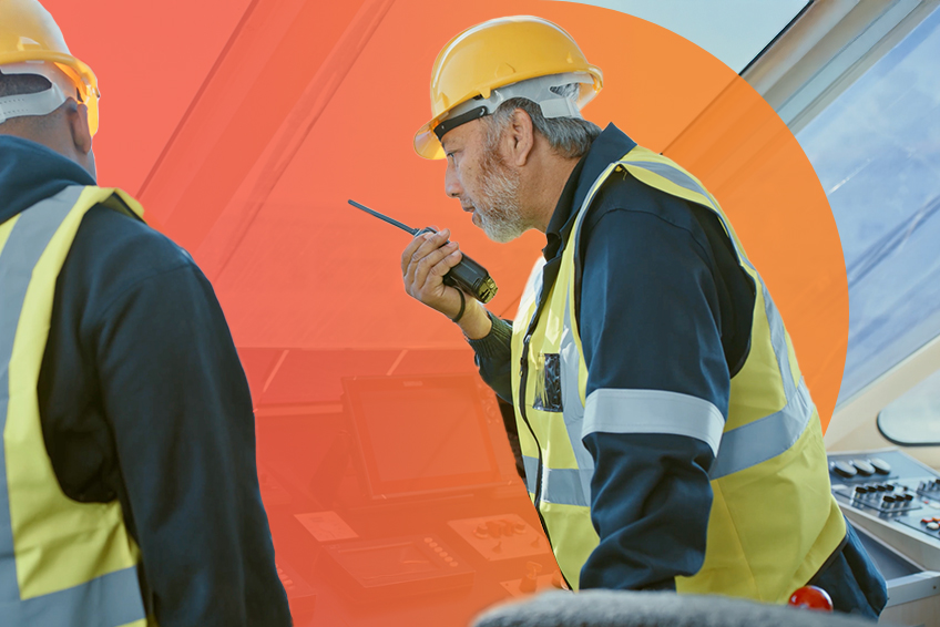 An individual wearing a reflective vest and hard hat speaks into a handheld radio.