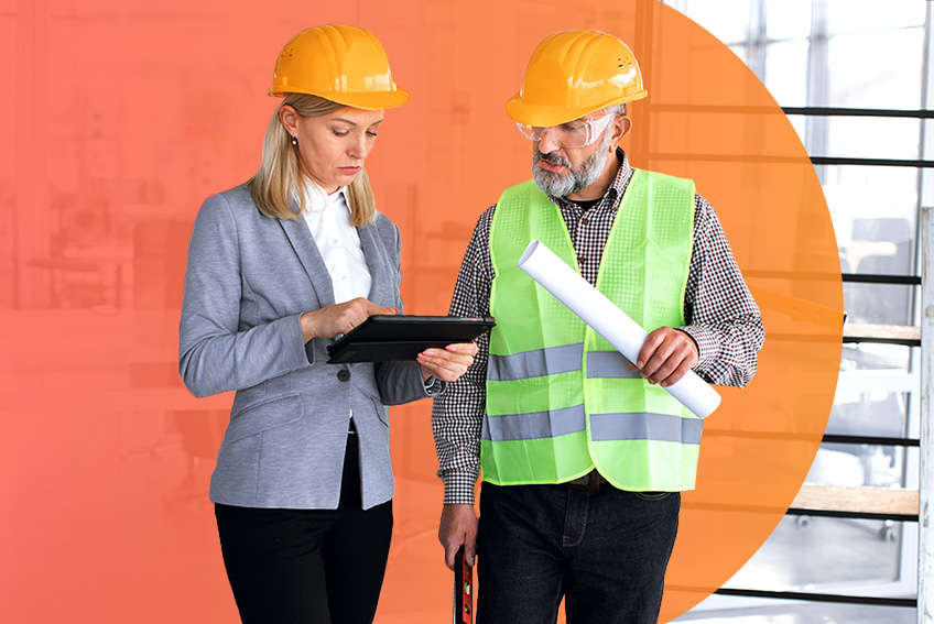 Two construction professionals wearing yellow hard hats are standing indoors discussing building plans. One person is dressed in a gray blazer and holding a tablet, while the other is wearing a neon green safety vest and holding rolled-up blueprints.