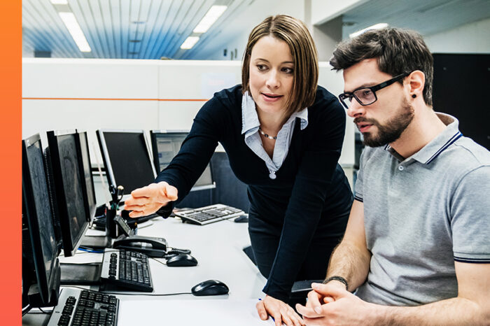 Two colleagues in a meeting room looking at computer screens and discussing work.