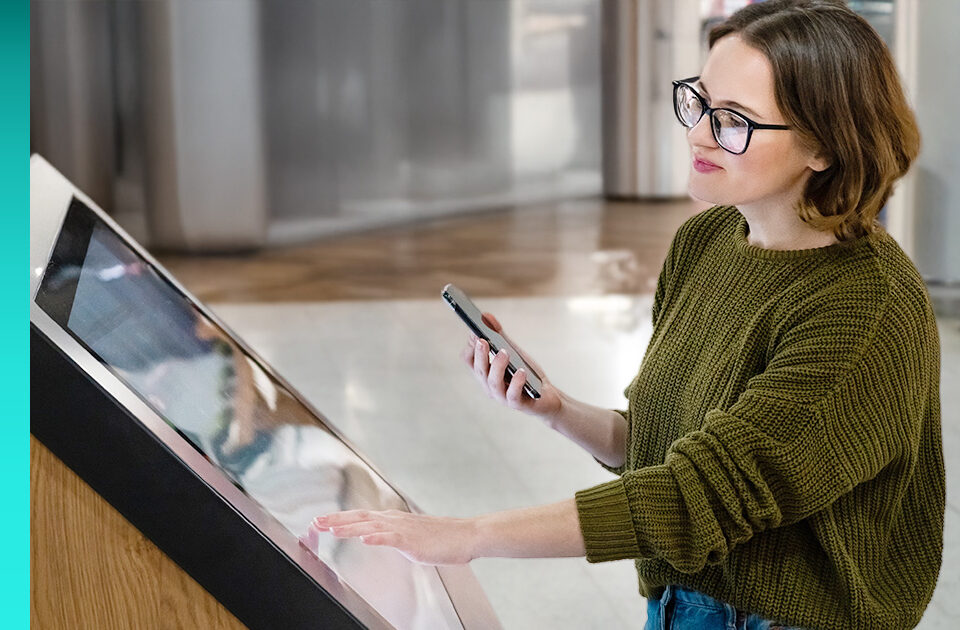 A person in glasses and a sweater holds a phone and interacts with a large tablet.