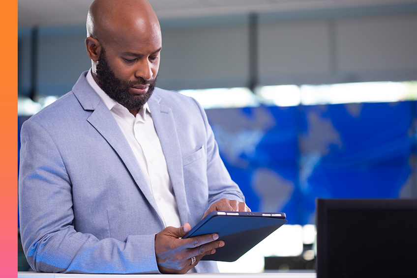 Businessperson in a light blue suit jacket and white shirt holds a tablet in a modern office with a world map display in the background.