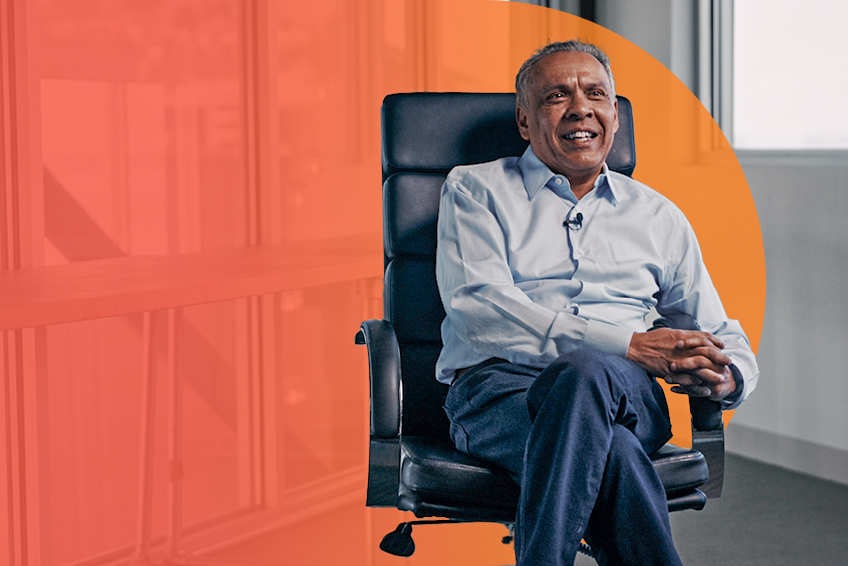 A person in a light blue shirt and dark pants sits in a black executive chair, smiling and looking to the left with hands clasped.