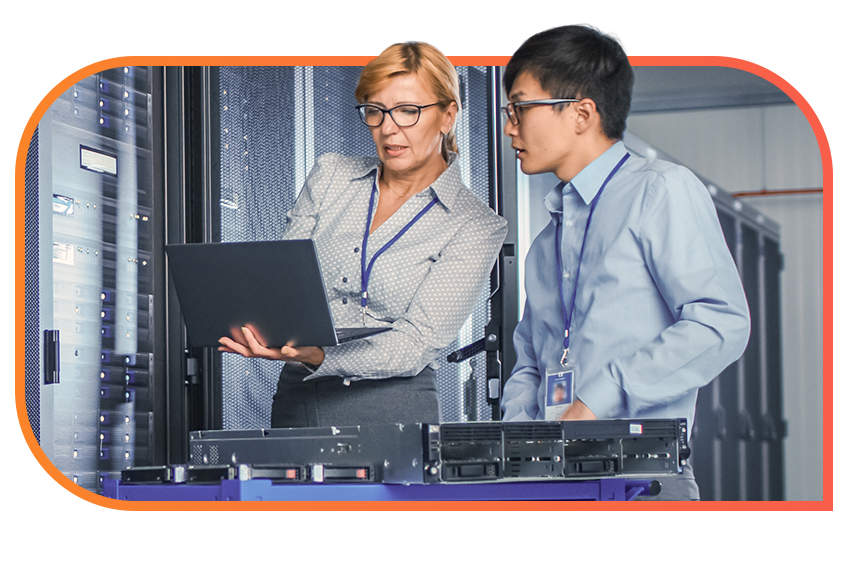 Two data center professionals are standing in a server room, one holding a laptop and showing something on the screen to the other, with racks of servers and hardware behind them.