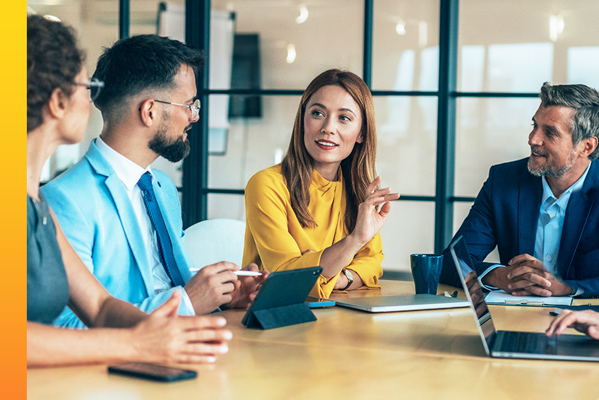 Four professionals in business attire sit around a table discussing how to deliver exceptional customer experience with laptops and a tablet in a bright office setting.