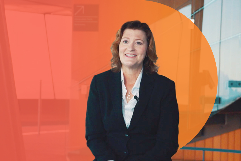 Smiling middle-aged businesswoman in a dark suit sits against a vibrant orange and red gradient background in a modern indoor setting.