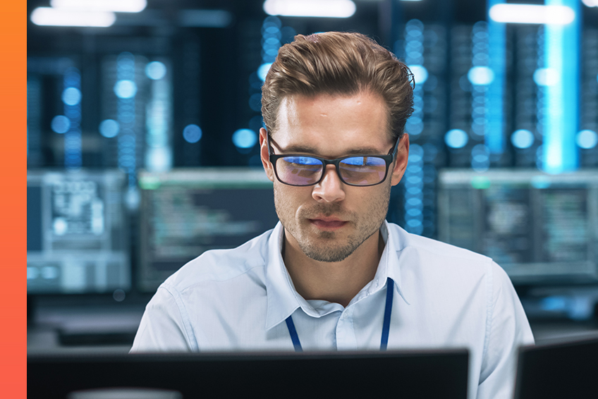 A professional with black glasses sits in with a blurred data center in the background, looking down at computer screens to work