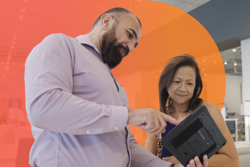 Two individuals in a furniture store stand together and look at a tablet one of them is holding.