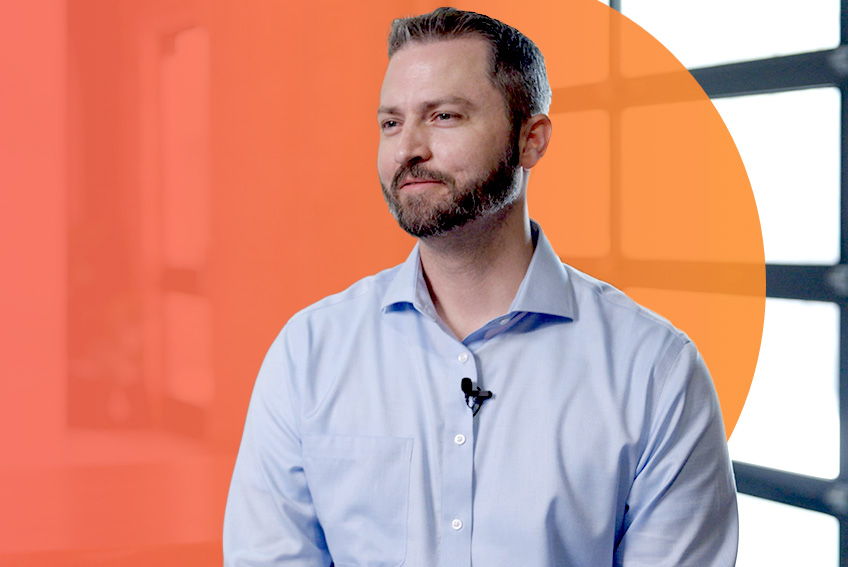 Person in a light blue button-down shirt stands in front of an orange gradient laid over windows.