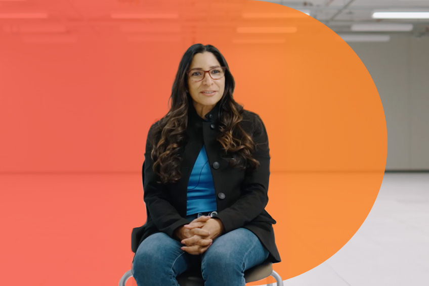 Person seated on a chair in a modern indoor setting with a gradient orange background, wearing a dark jacket over a blue top and jeans with hands clasped.