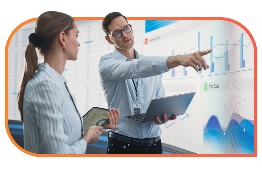 Two individuals holding tablets stand in front of a large screen with graphs, while one of them points at the screen and the other listens.