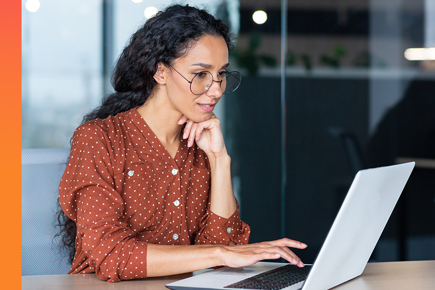 A person wearing glasses types on a laptop while resting their chin on their hand in a modern office setting.