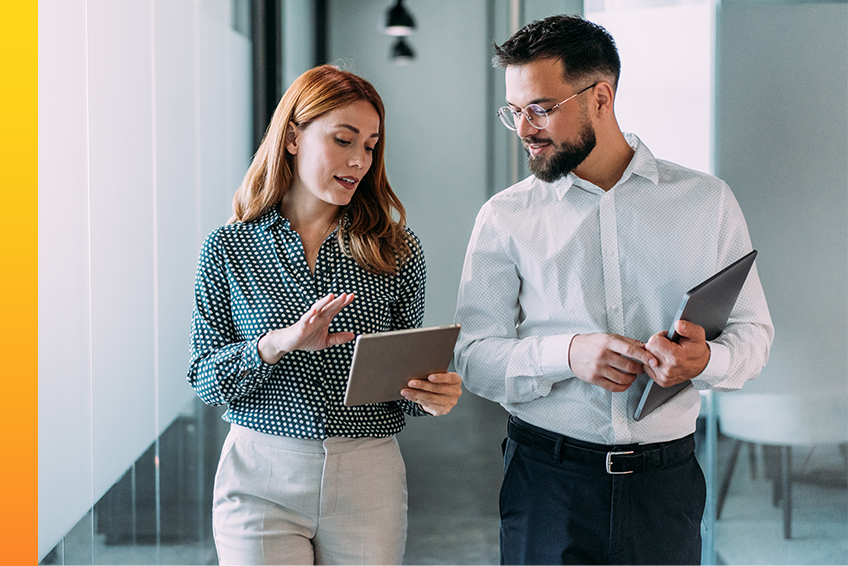A person carrying a tablet talks to a person with a laptop in a bright office.