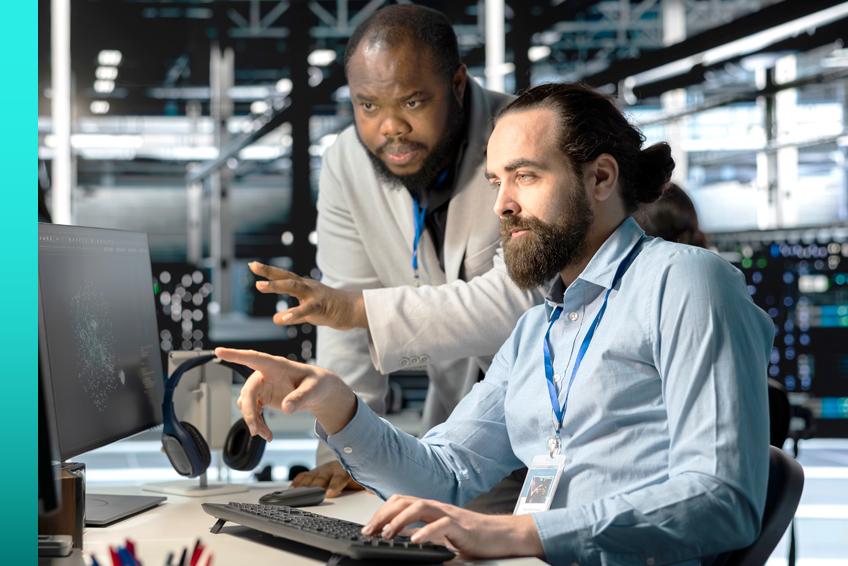 Two individuals in a modern office environment collaborating at a computer workstation. One person is seated, pointing at the monitor, while the other stands nearby, gesturing toward the screen.