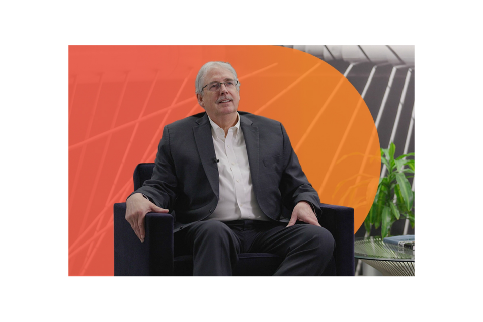 Robert J. McDermott seated in a modern armchair during an interview or discussion, wearing business attire, with a bold orange graphic backdrop and a potted plant nearby.