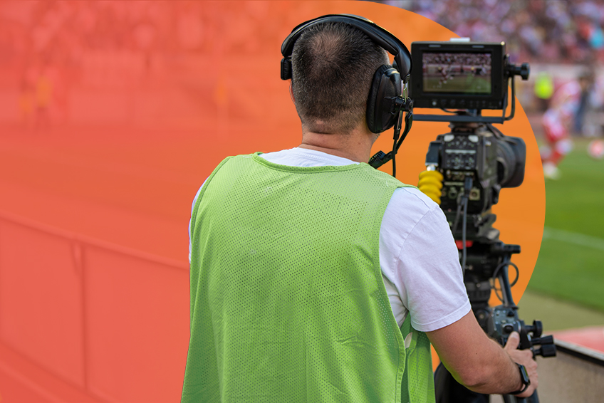 A cameraman in a green vest films a soccer field, with a digitally imposed orange gradient in the background.